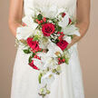 Held at the bride's waist like a precious keepsake, this cascading wedding bouquet captures a moment of quiet anticipation before the vows, the kind of scene you might glimpse outside a North Watford church on a sunny afternoon. The arrangement is dominated by sumptuous crimson red roses, each bloom full and velvety, spiralling inward with a depth that speaks of enduring love and devotion. Among them, pristine white lilies unfold, their wide, graceful petals and pale green centers offering a luminous contrast, as if brushed with morning light. Flowing from the heart of the bouquet, long stems of white Phalaenopsis orchids drift downward in a soft waterfall, their smooth petals and tiny buds adding rhythm and movement, like fabric cascading from the bride's hands. Tiny stars of white baby's breath weave in and out, softening the edges and creating a gentle, airy mist. Bright red berries, similar to hypericum, are tucked thoughtfully between the blooms, their glossy surfaces and rounded shapes adding playful texture and a hint of festivity, reminiscent of vibrant local floristry stalls near St Albans Road. Layers of rich greenery, from slender fronds to broad leaves, cradle the flowers and highlight their colours, giving the bouquet structure and a freshly gathered feel. The bride's white gown, with its delicate lace or embroidery, frames the arrangement without distraction, while a neutral cream backdrop keeps the focus entirely on the flowers. Designed in a classic red-and-white palette, this bouquet embodies romance and celebration, an ideal image for showcasing North Watford wedding flowers, cascading bouquet styles, or heartfelt, traditional bridal design.
