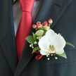 An intimate, detail-rich view of formalwear styling: the left lapel of a dark charcoal suit holds a single, immaculate Phalaenopsis orchid, its wide white petals soft and luminous under gentle indoor light. The orchid's lip carries a subtle yellow-gold pattern that punctuates the bloom's center with quiet elegance. Around the flower, deep green glossy leaves provide structure and a cool contrast in texture, while tiny clusters of baby's breath float through the arrangement like scattered confetti, lending a romantic, airy softness. A compact cluster of pink-to-coral hypericum berries sits nearby, their rounded forms and matte sheen picking up the warm tones of the scene and mirroring the rich satin red tie behind the boutonnière. The tie's sharp knot and the cuff of a light, subtly checked dress shirt add sartorial precision; a slim loop of red satin ribbon is tied at the base of the stems, a finishing touch that mirrors the tie and ties the composition together. I imagine this piece being worn by a groom stepping out near Watford Junction for photos, or by a guest arriving for a ceremony close to Vicarage Road; it's the sort of thoughtfully constructed boutonnière we prepare at Flower Delivery North Watford, combining tactile contrasts and coordinated colour so every detail reads with clarity and care on the day.