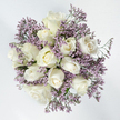 This image shows a circular bouquet photographed from directly above, resting on a bright, pure white background that echoes the calm mood of the flowers themselves. The heart of the arrangement is formed by around a dozen velvety white roses, each at a slightly different stage of opening. Some blooms are tight and modest, their outer petals just beginning to ease apart, while others are more fully unfurled, revealing richly layered centres that draw the eye inward. The roses glow with a creamy white tone, touched here and there by faint ivory shadows and a whisper of pale buttery yellow in the deepest petals, suggesting both innocence and warmth. Woven around and between these focal blooms are airy clusters of lavender-purple Limonium, whose many tiny blossoms create a soft haze of colour and a fine, delicate texture. This statice filler introduces gentle movement and depth, while its cool lilac shade beautifully offsets the warmth of the roses, giving the arrangement a composed yet tender character. Sparse hints of green stems and minimal foliage peek through, just enough to ground the bouquet in nature without disturbing the simple, two-colour harmony. The overall effect is one of quiet reassurance, like a soft-spoken message of sympathy. As a florist creating tributes for North Watford families, I think of this design as a comforting companion for condolence visits or memorial services, whether delivered to a home near St Albans Road or a small chapel nearby, where its understated elegance can offer a subtle sense of peace.