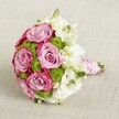 A luxurious hand-tied bridal bouquet rests on a softly textured, light beige fabric, viewed from a gently elevated angle that shows every curve of the flowers. At the centre, large, fully opened roses glow in romantic lavender-pink, their satin-like petals deepening to a richer fuchsia-pink along the outer edges, creating a graceful two-tone swirl. Nestled between the roses are bright chartreuse green button chrysanthemums, perfectly domed and neat, adding a crisp, modern pop of colour and playful texture. Delicate white blooms, likely lisianthus, weave through the design, their layered, ruffled petals and pale greenish-yellow centres softening the palette and giving the bouquet an airy, timeless elegance. A few tight green buds are scattered among the open flowers, hinting at just-cut freshness, as though picked that morning from a local grower on the outskirts of North Watford. The stems are gathered tightly into a smooth, comfortable handle, meticulously wrapped in glossy pink satin ribbon. Over the ribbon, a slim strand of tiny white pearls spirals down the length of the handle, catching the light like heirloom jewellery and evoking vintage romance. The neutral background lets the pinks, whites, and vivid greens shine, making the bouquet feel like it's glowing in gentle natural light. This arrangement would suit a bride walking through Cassiobury Park for photos, an elegant registry office ceremony near Watford town centre, or a special anniversary delivery in North Watford, where its blend of colour and texture speaks of love, celebration, and refined floral craftsmanship.