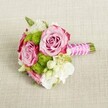 A small yet luxuriously detailed hand-tied bouquet lies horizontally on a softly textured light tan fabric, the weave suggesting natural linen on a table in a North Watford home. At the heart of the arrangement, three full, open pink roses draw the eye, their petals curving in delicate spirals from tightly furled centers to broader outer edges. Each rose shows a gentle ombre of colour, with blush pink deepening into a richer magenta along the tips, giving the flowers a painterly, romantic feel. Nestled between the roses are rounded tufts of lime green button chrysanthemums, their many tiny florets forming playful pom-pom spheres that add structure and contrast. Threaded through the bouquet are clusters of soft white and cream flowers, likely lisianthus or freesia, with thin, silky petals that bring airiness and a sense of calm. Tiny star-shaped white blossoms with yellow centers dot the gaps, like wax flower, creating intricate points of light against the more generous blooms. Subtle green foliage peeks out at the edges, grounding the design with fresh, natural tones. The stems are gathered into a compact handle, wrapped in a vivid fuchsia satin ribbon that glows against the neutral background. A strand of small white faux pearls winds diagonally around the ribbon, adding bridal elegance and a hint of vintage glamour. The neatly trimmed stems extend just beyond the ribbon, showing clean green ends. Soft, even lighting enhances the bouquet's pink, green, and white palette without harsh shadows, suggesting a calm indoor setting. The overall mood is romantic and polished, ideal for a bridesmaid's posy or intimate bridal bouquet carried at a wedding near Cassiobury Park or along St Albans Road in North Watford, where understated sophistication and freshness are cherished for special celebrations.