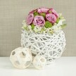 On a pristine white table, a rounded centerpiece glows in soft, even light, as if waiting to be placed along a wedding banquet in North Watford. The arrangement is anchored in a striking, spherical vase made of white, twig-like rattan strands woven into an open, basket-style globe, its airy gaps and curved lines giving an effortlessly contemporary feel while still echoing natural craftsmanship. Rising from this sculptural base, a tight dome of flowers unfolds: large pink roses and gentle lavender roses dominate, their petals layered and velvety, some with slightly deeper pink edges that create a sense of depth and intimacy. Between the roses sit bright green button chrysanthemums, neat and spherical, their vivid color injecting a burst of freshness that suggests early spring in nearby parks. Along the lower edge, small white filler blooms cluster and spill just slightly over the rim, softening the silhouette and adding a feathery lightness, as though the bouquet has been lovingly coaxed into perfect balance by an experienced local florist. To the left of the vase rest two decorative wicker spheres, one larger, one smaller, in pale natural tones that echo the woven motif and introduce a subtle sense of movement and rhythm. Behind the scene, a softly blurred, beige, linen-textured background creates warmth without pulling the eye away from the flowers, making the pastel roses and bright greens appear even more vivid. The composition feels intimate and carefully considered, ideal as a table arrangement for a North Watford wedding breakfast or a stylish engagement dinner at home.