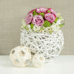 On a smooth white tabletop that gently mirrors every curve and petal, a romantic floral centerpiece glows as if prepared for an elegant North Watford wedding breakfast near St Albans Road. At its heart is a sculptural, spherical vase made from white-washed, interwoven twigs, the branches forming a light, open cage that cradles the flowers like a bird's nest redesigned for a contemporary home. Inside, a perfectly rounded dome of pastel pink roses, some tinged with deeper pink along their ruffled edges, opens softly towards the light, each bloom full and velvety. Colorful contrast comes from clusters of lime green button chrysanthemums tucked between the roses, their small, rounded heads giving a fresh, garden-like feel that brightens the whole piece. At the base, a gentle collar of creamy white blossoms-possibly lisianthus or similar dainty flowers-spills just slightly over the rim, their petals and buds blurring the line between the airy container and the lush bouquet. To one side on the table sit two pale wooden decorative spheres in an open lattice pattern, echoing the rustic weave of the vase and creating a calm balance in the composition, like carefully chosen ornaments in a well-loved North Watford living room. The background is a softly blurred wall in warm taupe, textured like woven fabric, which enhances the sense of depth and serenity. The entire arrangement radiates refined joy and tranquil beauty, ideal for understated wedding décor, civil ceremonies, or intimate anniversary dinners hosted across North Watford.