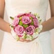 In soft North Watford morning light, a bride's delicate hands cradle a bouquet where dusty rose and mauve roses unfurl their velvety petals, each bloom suffused with a gentle touch of fuchsia at the tips. Between these romantic roses nestle button chrysanthemums in vibrant lime-green, their rounded forms and plush textures breathing fresh life and local energy-echoing the spirit found on nearby Exchange Road or Woodford Road. Encircling the bouquet, clusters of creamy-white spray roses or freesia form a gentle halo, a symbol of purity and new beginnings, with tiny green buds adding a sense of organic vitality. The bouquet's shape is classic and artfully balanced, perfectly complementing the bride's intricately patterned lace gown visible in the background. Her nails, elegantly painted pale pink with a hint of shimmer, mirror the delicate, romantic mood of the arrangement. Each element in this Watford-inspired bridal bouquet is thoughtfully chosen, blending soft pastels and lively greens to capture the promise of forever. The arrangement radiates tenderness and grace, ideal for a heartfelt spring or summer wedding along the leafy lanes of North Watford, and evokes a moment of gentle celebration deeply rooted in the local community.