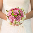 Held close against the intricate lace of a bridal gown, this bouquet embodies the gentle, heartfelt mood of a North Watford wedding morning. The eye is immediately drawn to the cluster of lush lavender-pink roses at the centre, each rose opening in generous, velvety layers that suggest both elegance and warmth. Between them, bright lime-green button chrysanthemums peek out playfully, their rounded, slightly textured petals catching the light and providing a modern jolt of colour that makes the roses seem even softer by contrast. Encircling the arrangement, small creamy-white blooms and slender buds create a delicate halo, almost like a ring of floral lace that mirrors the detail on the bride's dress. The bouquet is hand-tied, stems neatly gathered so it rests naturally in her hands, ready to be carried down an aisle in a local North Watford church or civil venue. In the mind, a subtle scent rises: the familiar perfume of fresh roses mixed with the cool, green aroma of newly cut stems, reminiscent of an early stroll past gardens near Garston or around Leavesden. The light in the image is gentle and flattering, catching the curves of each petal and the bright green centres of the chrysanthemums, giving the bouquet a quietly radiant glow. As a florist used to preparing wedding flowers that last from first look to evening reception, I can almost feel the resilience of these blooms, designed to remain composed through hugs, photographs, and celebration. The bouquet speaks softly of joy, new beginnings, and the comforting beauty of flowers gathered with care in North Watford.