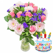 A bright, celebratory bouquet sits at the centre of a clean white background, the kind of arrangement a neighbour might bring from a local Watford florist to mark a birthday. The composition is dominated by soft pink roses-petals unfurling from blush to a deeper pink-paired with pale pink Gerbera daisies whose broad, cheerful faces show buttery-yellow centres. Sprays of vivid purple filler flowers, likely statice or limonium, are tucked through the blooms, their papery texture creating a lively contrast against glossy deep-green foliage that frames and lifts each blossom. The stems sit in a simple clear glass vase with water visible at the base, the transparency emphasising freshness and careful craftsmanship. To the lower right, a cake-shaped foil balloon adds a playful note: a white 'frosted' surface edged with wavy blue icing, scattered sprinkles in red, yellow, blue and green, and four striped candles-red, yellow, green and blue-topped with tiny yellow flame motifs. The words 'Happy Birthday' arch across the front in bright, multi-coloured letters, while a small 'KALEIDOSCOPE' mark appears discreetly at the balloon's base.

Soft morning light kisses the petals and makes the varnished leaves gleam; the scent is a gentle, sweet whisper rather than overpowering perfume. As a florist who often delivers into North Watford-from Nascot Wood lanes to the bustle around Watford High Street-I picture this arrangement arriving with a handwritten tag, a small, festive surprise that pairs classic floral romance with a cheeky party accent for someone's special day.