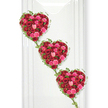 Three rose-filled hearts descend in a graceful line, forming a poignant and romantic funeral tribute that feels both modern and timeless. Arranged against a smooth, glowing white background, the hearts appear to float in space, each one brimming with densely packed roses in shades of deep crimson, vivid fuchsia, and tender blush pink. The intense red blooms provide a foundation of unwavering love and respect, while the pinks soften the composition with notes of compassion, warmth, and fond remembrance. Every heart is meticulously shaped, the roses set so closely that their velvety petals overlap, creating a luxurious surface that invites the eye to wander across each curve. Some flowers are tight buds, filled with potential, while others are fully open, symbolising a life fully lived and shared. Delicate ivy vines in rich, glossy green trace the outer edges of every heart, adding an organic frame that feels almost like an embrace. These slender tendrils stretch down between the hearts, gently linking them into one continuous chain of affection and memory. Soft, even lighting ensures the colours appear vivid and true, and the white backdrop keeps the focus entirely on the floral artistry without any visual clutter. This style of tribute would be chosen by families in North Watford who wish to honour a loved one with an arrangement that speaks eloquently of deep connection, whether placed near the coffin, beside an urn, or close to a framed photograph during a memorial service or gathering at home.