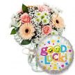 A bright, encouraging gift set captured against a clean white backdrop: a generous glass vase tied with a simple twine bow holds a lush, hand-styled bouquet of soft peach and light pink roses and warm peachy-orange Gerbera daisies with their deep brown centers. Tiny white gypsophila clouds and small white-and-lime-green chrysanthemum florets are sprinkled through the composition, while rich dark green foliage peeks between blooms to give weight and depth. The roses' velvety petals and the Gerberas' satiny faces catch soft studio light, suggesting the faint, familiar perfume of roses balanced by the crisp, green note of chrysanthemum. To the right, slightly overlapping the lower vase, a round silver metallic foil balloon declares Good Luck! in bouncy, multi-coloured letters; two yellow smiley faces replace the o's, one winking and one sticking its tongue out, and playful hearts, stars and confetti shapes in blue, pink, yellow and green scatter across the silver sheen. A small KALEIDOSCOPE logo sits near the balloon's bottom edge. The whole arrangement reads as a tactile, optimistic gesture - the rustic twine and clear glass make it feel handcrafted, while the cheerful balloon adds celebratory sparkle. As a florist who often delivers to nearby Cassiobury Park picnics and to neighbours around Oxhey and Watford High Street, I picture this set arriving for exam send-offs, interview send-offs, graduations or a warm farewell - a simple, fresh composition meant to lift spirits and send unmistakable best wishes in North Watford.