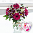 A richly textured floral display is shown resting on a clean, light-coloured surface, glowing in gentle daylight like a surprise delivery arriving at a North Watford doorstep. The bouquet features several proud gerbera daisies in intense fuchsia and magenta, their wide petals radiating around dark, almost ink-black centres that ground the arrangement. Around these focal blooms cluster intricate chrysanthemums, each small flower made up of layered petals that shift from deep purple to snowy white at the tips, encircling warm yellow centres that sparkle like tiny suns. Creamy white roses weave between the stronger colours, their softly rolled petals adding a sense of grace and quiet romance. Airy sprigs of white baby's breath scatter through the bouquet, bringing a delicate lace-like lightness that softens the richer tones. The design is completed with fresh, green foliage that peeks out from beneath, shaping the bouquet into a generous, rounded form that feels full but never crowded. All of the blooms are gathered in a clear glass vase, its simplicity allowing the neatly trimmed green stems and clear water to be seen, suggesting freshness and professional care. Around the neck of the vase, a piece of rustic natural twine is tied in a casual bow, suspending a small white gift tag featuring a stylised pink rose motif and green handwritten-style wording that makes the arrangement feel personal. Just to the right, a shiny heart-shaped foil balloon with a reflective silver border adds a playful, loving touch. Its white centre carries the phrase Love n' Hugs 24-7 in a collage of patterned letters in pinks, purples, and blues. The softly blurred, bright background suggests an airy indoor space near a window, capturing the mood of a thoughtful, affectionate gift for birthdays, romantic surprises, or simply sending love across North Watford.