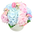 A generous dome of pastel flowers rises from a sleek white hatbox, captured against a spotless white background so that the colours appear especially soft and luminous. At the base of the composition, large, rounded heads of light blue hydrangea create a cool, cloudlike foundation, each floret contributing to a plush, almost cushiony texture. Nestled into this blue are voluptuous pink peonies, some tightly layered and others blooming wide open, their ruffled petals spilling gently outward and offering a sense of classic romance. Threaded among them, smaller blush-pink spray roses peek out in various stages of bud and bloom, providing intricate detail and a scattering of tiny focal points that invite the eye to wander. Two striking hot-pink calla lilies rise with graceful curves from the arrangement-one positioned towards the upper left, the other on the right-adding elegance and a slightly more vivid note that keeps the design feeling current and sophisticated. A few deep green leaves are barely visible at the base, offering a subtle natural contrast without disturbing the pastel serenity. The round white hatbox, with its understated ridge near the top, acts as a minimalist pedestal, almost concealed by the sheer abundance of blossoms that seem ready to spill over the edge. The overall feel is like a quiet celebration, something you might spot on a windowsill overlooking North Watford's residential streets, bringing softness to a modern flat or traditional home. It is easy to imagine this arrangement marking a new baby's arrival, a gentle Mother's Day surprise, or a message of calm support during a difficult time, delivered fresh and full of care within the local community.