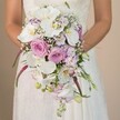 In this evocative North Watford wedding scene, a close-up eye-level portrait unveils a bride in mid-torso, her delicate hands holding a luxurious, teardrop-shaped bridal bouquet. The focus falls on a cascade of pristine white Phalaenopsis orchids, their broad petals gleaming beneath soft, natural light, their centers glowing with subtle yellow gold. Surrounding the orchids are lush roses in pastel shades of pink, blush, and lavender-some just beginning to open, others fully blossomed, their velvety layers recalling the fresh scents of Watford's Saturday flower markets. Wisps of white baby's breath float amongst the larger blooms, lending an ethereal, cloud-like softness and inviting the touch of spring air. Scattered within are violet and lavender blooms, perhaps sweet pea or fragrant stocks, contributing gentle fragrance and extra depth. Cascading strands of richly hued fuchsia Amaranthus add a dramatic, flowing accent, their unique textures providing contrast and modern flair against the romantically classic composition. Gentle sprays of verdant green foliage and trailing buds peek through, reflecting the productive gardens and abundant greenery found around North Watford. The bridal gown, with subtle lace detailing and a sweep of smooth, white fabric, offers refined elegance, setting the bouquet centre stage against a calm, muted background. This arrangement encapsulates both tradition and contemporary grace, making it an inspiring choice for North Watford couples dreaming of a memorable, sophisticated spring or summer ceremony.