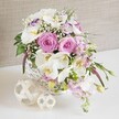 A lush, romantic table arrangement of pink and white flowers rests in a round white wicker basket, styled as if ready to adorn a wedding table in North Watford. At its heart are abundant pink roses, fully open and velvety, their spiralled petals catching the light and suggesting warmth, affection, and timeless elegance. Among them, white Phalaenopsis orchids rise and curve, each bloom with broad, smooth petals and a golden-yellow throat that adds a sophisticated, almost sculptural accent. Frilled white Lisianthus, some kissed with a subtle lavender edge, nestle between the larger blooms, while slender stems of soft pink stock extend outward and slightly downward, giving a sense of gentle cascade. Airy clusters of white baby's breath weave through the bouquet like delicate lace, softening spaces and lending a dreamy, cloud-like effect. Rich green foliage peeks from beneath and around the flowers, providing contrast and a sense of freshness, as though the arrangement has just been composed for same-day delivery to a nearby North Watford hotel or family home. The white wicker basket, with its intricate weave, adds rustic charm and a hand-crafted feel, and it stands on a pristine white tabletop that highlights every colour and curve. To one side rest two woven decorative spheres in pale natural tones, their open, interlaced strands echoing the basket's texture and adding a contemporary decorative note. Behind, a soft beige fabric backdrop, subtly textured, creates a warm, neutral setting that allows the pinks and whites to glow. The scene conveys relaxed luxury and joyful celebration, ideal for wedding receptions, anniversary dinners, or elegant parties across North Watford.