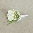 A single white orchid boutonniere rests gently on a softly textured, light beige woven fabric, the kind of natural linen you might find in a calm North Watford townhouse near Cassiobury Park. At the heart of the design sits a flawless Phalaenopsis orchid bloom, its broad, velvety petals a clean, luminous white that curves outward in a quiet, sculptural arc. In the centre, the orchid's throat reveals a faint blush of buttery yellow and the softest hint of cream, adding warmth and a touch of depth to its otherwise serene palette. Surrounding the bloom, countless tiny white baby's breath flowers float like a cloud, each minuscule floret forming a halo of softness that breaks up the smooth lines of the orchid and gives the boutonniere a romantic, airy feel. Threaded between these delicate gypsophila clusters are fine strands of feathery green foliage, similar to asparagus fern or a very light maidenhair, their fresh green tones suggesting the scent of a summer garden after light rain over North Watford's residential streets. The stems are neatly gathered at the base and wrapped in a pristine white satin ribbon, finished with a small, carefully formed bow that looks ready to be pinned to the lapel of a groom or groomsman. The lighting is gentle and even, without harsh shadows, so every texture - the matte fabric background, the glossy orchid petals, the frothy baby's breath, and the smooth ribbon - can almost be felt through the description. The overall impression is of understated elegance and timeless purity, the kind of floral detail chosen for a wedding morning that begins quietly at home before heading to a ceremony in one of the local churches around North Watford.