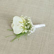 A refined white orchid boutonnière rests at a slight angle on a pale tan, linen-like fabric, its understated design capturing the essence of a modern North Watford wedding. At the heart of the piece is a single, perfectly formed orchid bloom, its petals wide and gently curved, as smooth and luminous as fine porcelain. The petals are a cool, crisp white, gathering toward a centre touched with the softest hint of buttery cream and pale yellow, adding warmth and dimension without overpowering the simplicity of the design. Encircling the orchid are wisps of white baby's breath, the numerous tiny blossoms creating a soft, cloud-like frame that brings lightness and an almost romantic haze to the composition. Delicate strands of bright green foliage slip from beneath the flowers, their thin blades arching gracefully and giving a subtle sense of movement, like a breeze passing through the greenery along St Albans Road. The stems are neatly aligned and expertly wrapped in smooth white satin ribbon, creating a polished handle that feels both comfortable and secure for pinning to a lapel. A small satin bow, tied just below the blooms, offers a classic finishing touch, suggesting careful craftsmanship and attention to detail. The background fabric has a gentle texture and natural weave, adding depth and a quiet, organic feel, much like a well-tailored suit laid out before a ceremony. Soft, diffused lighting washes over the boutonnière, highlighting the different textures-the sleek orchid, the airy baby's breath, the crisp greenery, and the lustrous ribbon-evoking a mood of calm sophistication, perfect for a groom or honoured guest arriving at a local venue for a day of celebration and new beginnings.