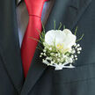 Tucked into the top pocket of a tailored charcoal jacket, this elegant boutonnière looks ready for a wedding celebration somewhere between North Watford and the tree-lined streets near St Albans Road. The focal point is a single white orchid, its petals smooth and curved like sculpted porcelain, catching the light with a soft, creamy glow. At its centre, a subtle golden hue glimmers, a tiny heart of colour that adds depth and warmth. Surrounding the orchid, a gentle spray of baby's breath creates a dreamy halo, each miniature flower contributing to an airy, cloud-like effect. Slender stems of fresh greenery and fine, feathery fronds weave through the design, bringing a lively, natural rhythm that contrasts beautifully with the structured lines of the suit. At the base, a slim white satin ribbon is tied with precision, gathering the stems into a compact, polished finish that speaks of skilled craftsmanship. A bright red tie runs down the middle of the image, its vivid shade echoing the joy and passion of a wedding day, and making the white blooms seem even more pristine. The overall mood is one of understated romance and timeless style, as if the wearer is just about to step into a registry office ceremony or arrive at a North Watford reception venue, carrying with them the soft scent and sincere emotion of thoughtfully chosen flowers.