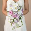 Against a muted, gently diffused tan backdrop, mirroring North Watford's classic charm, a bride in a strapless white gown stands waist-up, triumphantly holding a lavish cascading bouquet crafted for her wedding day. The flowers create a poetic arc: generous white orchids with yellow and violet-blushed centers mark the arrangement with polish and grace, standing above pillowy pink garden roses whose delicate fragrance drifts in the air, evoking leisurely walks through Watford's springtime parks. Smaller lavender and pale violet blossoms nestle between the larger flowers, their feathery shapes adding depth and subtleness. Peppered throughout are sprigs of baby's breath, frothy and bright, with sparse green leaves emerging like the soft undergrowth below a woodland canopy. The intricate lace patterns along the bride's dress peek through just below the bouquet, underscoring a look of refined daring and romance. The bouquet's sweeping silhouette is both classic and modern-a composition that would grace both stately hotel receptions and intimate garden ceremonies alike in North Watford. Through professional artistry and understanding of the community's taste, this arrangement showcases how bespoke bridal flowers can become the heart of local celebrations, bringing together enduring sophistication and the fleeting beauty of each bloom in one unforgettable North Watford wedding keepsake.