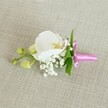 A delicate white orchid boutonniere is laid horizontally on a pale oatmeal linen backdrop, its soft, woven texture giving the scene a calm, understated elegance that feels right at home in a North Watford townhouse or registry office. At the heart of the design, a single Phalaenopsis orchid bloom opens in pristine white, its broad, velvety petals curving gently around a centre touched with pale yellow and the faintest whisper of pink. The flower looks freshly picked, as if prepared early in the morning for a groom or prom guest heading out from Garston or along St Albans Road. Around the base of the orchid, tiny tufts of white baby's breath cluster like light clouds, each miniature blossom adding a dreamy, airy volume that softens the lines of the design. Sprigs of slender, bright green foliage peek out from behind, bringing a lively contrast to the white petals and echoing the green spaces of parks around North Watford. To one side, a short stem extends, tipped with smooth, lime-green orchid buds that are still tightly closed, suggesting quiet anticipation and the promise of future moments. The stems are carefully bound with a narrow fuchsia pink satin ribbon, tied into a neat, refined bow; its glossy surface catches the light and introduces a bright, celebratory note against the neutral fabric. The gentle, even lighting enhances every texture-the satin sheen, the linen's subtle weave, the orchid's satin-like petals-creating an impression of calm preparation before a joyful event such as a wedding, civil ceremony, or school leavers' prom in the local area.