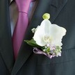 A close-up photograph shows a sharply tailored charcoal suit jacket, the left lapel filling the frame like a dark, elegant backdrop for a single white orchid boutonniere. The fabric of the suit has a fine, almost matte weave, catching just enough light to reveal careful pressing and a clean edge, the kind you might see on a groom stepping out of a car near Cassiobury Park for a North Watford wedding. Nestled against this deep grey is a lustrous tie in rich magenta leaning softly towards plum, its smooth silk knot resting against a pale grey dress shirt woven with a discreet, textured pattern that adds quiet depth rather than shine. Pinned precisely to the lapel, the Phalaenopsis orchid is the star: broad, satin-like petals open generously, pure white with a sculpted curve that looks almost like folded porcelain. At the centre, a touch of warm yellow and soft orange glows around the stamen, bringing life and warmth to the cool palette. Around the orchid, petite lavender blossoms and tiny cloud-like tufts of white gypsophila cluster in gentle sprays, creating a halo of softness. Deep green leaves cradle the flowers, their glossy surfaces adding a fresh, just-cut feeling that suggests same-day preparation. Above the main bloom, a single pale green orchid bud rises, still closed, hinting at new beginnings. The stems are wrapped at the base with a neatly tied lavender satin ribbon bow, its subtle sheen echoing the tie's hue. The whole composition feels calm, refined, and romantic, the kind of understated boutonniere a North Watford florist would design for a thoughtful, modern celebration.