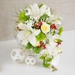 A beautifully composed cascading arrangement of white flowers is presented on a bright white tabletop against a soft, beige textured backdrop, evoking the calm, elegant atmosphere of a North Watford wedding breakfast or anniversary dinner. The design is anchored in a round white wicker basket whose woven structure adds gentle texture and an artisan touch. From this base, several large white lilies rise and open, their trumpet-shaped petals curving outward to reveal speckled throats and golden anthers, capturing both drama and purity. Nestled among the lilies, a single cream-yellow rose forms a warm focal point, its compact, spiralling petals offering a subtle contrast to the lilies' bold shapes. On the right, a graceful cascade of pure white Phalaenopsis orchids drapes downward in a soft arc, giving the arrangement a waterfall or teardrop silhouette typical of stylish bridal bouquets. Surrounding the central blooms is a lush halo of greenery: wispy grasses, feathery foliage, and rounded glossy leaves that add depth and frame the flowers. Bright green pom-pom style blooms, reminiscent of green trick or small chrysanthemums, tuck in between the foliage, bringing a vivid, moss-like texture. Fine sprays of white baby's breath weave delicately through the design, while clusters of glossy red berries supply vivid, festive dots of colour, like scattered rubies. To the left of the basket, two white woven spheres in different sizes rest on the table, their airy lattice echoing the basket's texture and completing the scene. The arrangement feels fresh, balanced, and thoughtfully crafted, the sort of centrepiece a local North Watford florist might prepare for intimate gatherings, civil ceremonies, or special family celebrations.