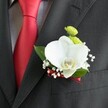 A close, intimate photograph focuses on a single wedding boutonniere pinned neatly to the lapel of a dark charcoal suit jacket, the finely woven wool catching the light in soft, diagonal threads. At the heart of the design is a flawless white Phalaenopsis orchid, its broad, velvety petals opening like wings around a delicate yellow-and-white lip that adds a gentle warmth to the centre. Tucked just above the orchid, two small, perfectly rounded lime-green chrysanthemum button blooms provide a fresh, modern contrast, their tiny petals forming tight, textured domes. Sprigs of white baby's breath weave around the main flowers, each miniature bloom like a speck of morning cloud, softening the edges of the composition. A few glossy, deep green leaves cradle the flowers and give the piece a grounded, natural frame. At the base, a narrow deep red satin ribbon is wrapped and secured with care, mirroring the rich red of the silk tie that lies to the left against a pale grey and white checked shirt. The tie's sheen and striking colour echo the romance of a wedding ceremony, perhaps in a North Watford registry office or a nearby church off St Albans Road. Light falls gently across the scene, creating subtle highlights on the orchid's curved petals and the crisp lapel, suggesting a moment of quiet anticipation before vows are spoken. The boutonniere feels refined yet personal, a thoughtfully crafted floral accent that adds a whisper of luxury and tenderness to a groom's attire on a day of love and celebration in the heart of the local community.
