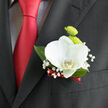 The image shows an elegant wedding boutonnière pinned to the lapel of a dark, formal suit, the kind of refined detail often seen at ceremonies around North Watford. At its heart, a single white orchid opens with sculptural grace, each petal thick and velvety, marked by a hint of buttery yellow at the centre that adds warmth to the cool white. Tucked just behind are a pair of lime-green button chrysanthemums, small and round, their intricate petal pattern forming a textured cushion that contrasts beautifully with the orchid's smooth surface. Wisps of baby's breath arc softly around the flowers, their tiny snow-white blossoms creating a halo effect that softens the edges of the design. Rich, glossy green leaves peek out from the back, grounding the composition and providing a deep backdrop against the black lapel. The stems are bound tightly with a narrow, deep red ribbon, its colour mirrored in the groom's satin tie that glows beneath, adding a thread of romance and passion to the otherwise crisp palette. The lighting is gentle, likely indoors at a hotel or reception venue not far from North Watford's high street, giving the flowers a luminous, almost dewy quality. This boutonnière feels intentionally understated yet full of meaning, perfect for a groom who prefers subtle luxury-something you can imagine being carefully prepared on the morning of the wedding, set aside in a cool box, and then pinned with steady hands just before the walk down the aisle.