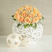A dome of honeyed peach roses, each bloom layered with velvety spirals, rests within a modern white rattan sphere, the arrangement radiating a gentle, celebratory hush. Tiny clusters of white gypsophila are woven through the roses like whispered vows, their airy texture scattering light and lending the bouquet a bridal softness that feels both tender and refined. Lush green leaves peek between the blooms, their cool tones grounding the warm apricot hues and adding depth to the rounded silhouette. The white woven vessel acts as a sculptural pedestal, its open-work pattern catching morning light that seems to gild the petals with a faint golden wash. Close your eyes and you can almost sense the soft, powdery fragrance rising from the roses, mingling with the crisp scent of greenery-a combination that evokes quiet walks by Watford High Street or a rehearsal dinner shaded by trees near North Watford station. Designed with the care of an experienced local florist, this arrangement suits small weddings, milestone anniversaries, or an elegant gift to mark a new beginning. Flower Delivery North Watford often prepares this style for customers seeking something contemporary yet classic, made to complement both modern homes and traditional venues. Every element is considered-the domed shape, the airy pockets of baby's breath, the tactile contrast between silk-like petals and woven rattan-creating a bouquet that looks as beautiful up close as it does from across the room, offering a serene, heartfelt presence.