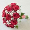 Centred against a softly lit, understated background, this bouquet of vibrant red roses and airy white baby's breath embodies elegance and joy-an arrangement expertly handcrafted by North Watford florists to suit the area's traditions of heartfelt celebration. The rounded, dome-like design highlights the velvety texture and rich, passionate hue of each rose in full bloom, some petals still curled tightly, while the interspersed clusters of gypsophila add a whisper-light, cloudlike quality. Subtle flashes of dark green foliage peek through, grounding the composition with a natural touch reminiscent of strolls through North Watford's green spaces. A wide red satin ribbon wraps the handle neatly, adorned with crisp white stripes and glimmering pearls that spiral gracefully, offering a tactile sense of romance and detail. The freshly cut stems peek out at the base, promising longevity and natural freshness ideal for a wedding bouquet, anniversary surprise, or cherished local delivery across North Watford's neighbourhoods. The overall effect is one of classic romance and celebration, echoing the symbolic meanings of enduring love and new beginnings that red roses and baby's breath hold for so many. Bathed in gentle, diffuse light, each detail-petal, leaf, and pearl-stands out beautifully, making this bouquet a timeless focal point for unforgettable moments in the heart of North Watford.