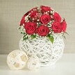 A beautifully composed floral scene showcases a rich, dome-shaped bouquet of red roses that immediately steals the eye, each bloom fully unfurled so the petals create velvety spirals, tight at the centre and softening elegantly towards the edges. The roses, in a deep and romantic crimson, are arranged closely together until they form a smooth, almost spherical crown that suggests the care of a professional florist preparing a wedding table in North Watford. Nestled between the larger blooms are filmy clusters of white baby's breath, their tiny blossoms scattered like stars across the arrangement, bringing a delicate, cloud-like quality that balances the intensity of the red. A few flashes of fresh green foliage peek from within, adding depth and a subtle reminder of the garden. The bouquet is anchored in a notable white vase, rounded and slightly bulbous, created from slender strands that weave and interlock into an airy lattice. This open-weave structure lets glimpses of the stems and the space behind shine through, giving the vase a crafted, contemporary feel that would sit comfortably on a reception table at a local North Watford venue or in a modern dining room. In front of the vase, resting on a creamy white surface, two pale woven spheres echo the same basket-like pattern, the larger orb placed slightly ahead of the smaller one to create visual rhythm. They feel like calm, sculptural accents, adding texture without distracting from the flowers. The backdrop is a softly blurred wall in warm, neutral tones, free of clutter, so every nuance of colour and weave stands out. The overall impression is of serene, romantic elegance, a centrepiece that quietly elevates any celebration.