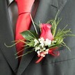 A single, thoughtfully arranged boutonniere is shown in close-up, pinned to the lapel of a dark charcoal grey suit, as if worn by a groom or groomsman at a North Watford wedding. At its centre rests a deep pink, almost fuchsia rose bud, partially opened so that its velvety petals curl softly inward, creating a sense of quiet romance. Around the rose, delicate clusters of white gypsophila form a light, airy collar, each tiny bloom giving the impression of a fine bridal veil. The greenery is carefully layered: glossy, broad leaves nestle close to the flower, while slender, feathery sprigs reach outward, adding height and texture without overpowering the design. At the base, a small, bright red satin ribbon is tied into a neat bow, its sheen echoing the rich red silk tie lying beneath the lapel. The suit fabric appears to be a high-quality wool blend in charcoal grey, with a subtle texture that catches the light, and a softly patterned grey dress shirt peeks out from under the jacket, adding gentle depth to the scene. The light in the image is soft and even, enhancing the colours of the rose and greenery while keeping the overall tone elegant and calm. It feels like the quiet moment just before walking down the aisle at a local venue near St Albans Road or along the streets of North Watford, a small but meaningful floral accent that conveys love, care, and a sense of occasion crafted by a skilled local florist.