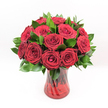 A luxurious, tightly clustered bouquet of deep crimson red roses fills the frame, each bloom perfectly formed and velvety, as if freshly gathered by an expert florist in North Watford. The roses range from buds just beginning to unfurl to fully open blossoms with layered petals spiralling outwards, creating a rich, plush dome of colour. Their intense ruby tones feel almost luminous against a collar of dark, glossy green foliage, likely Ruscus or similar leaves, which cradle and separate the blooms while adding depth and freshness. Every leaf catches the light with a subtle sheen, giving the arrangement a polished, well-tended appearance. The bouquet is nestled in a translucent ruby-red glass vase with a gently twisted, spiralled design running up its sides, the curves in the glass creating delicate reflections and highlights. Through the tinted glass, you can just make out the sturdy rose stems, arranged with care so they fan out evenly, providing a strong foundation. The vase has a slightly flared rim and a solid, reassuring base, anchoring the abundance of flowers above. The entire image is set against a pristine, white background, which intensifies the contrast between the vibrant red roses and the lush green foliage, making the arrangement appear almost three-dimensional. Bright, even lighting reveals every petal edge and leaf vein, suggesting flowers at their peak of freshness, like a premium bouquet ready for delivery to a doorstep near Cassiobury Park or along St Albans Road. The overall mood is romantic and celebratory, ideal for anniversaries, Valentine's gestures, or any heartfelt declaration of love and admiration in and around North Watford.