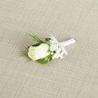 A classic single white rose boutonniere lies diagonally across a softly textured, light beige fabric that resembles finely woven linen, the kind you might see on an elegant table in a North Watford townhouse near Cassiobury Park. At the heart of the design is a creamy white rosebud, its velvety petals still gently furled, suggesting a bloom cut at peak freshness, as if prepared early that morning by a careful local florist. Around the rose, tiny clouds of white baby's breath form a delicate halo, each miniature blossom adding a lace-like softness that contrasts with the smoothness of the central flower. Slender, deep green leaves and a few feathery, fern-like sprigs cradle the rose, their rich colour intensifying the purity of the white petals and echoing the greenery of North Watford's quiet residential streets and gardens. The stems are neatly gathered and wrapped in a pristine white satin ribbon, pulled snug and even, and finished with a small, tidy bow that sits just beneath the rose, ready to be pinned precisely to a lapel for a wedding ceremony at a nearby church or registry office. The lighting is gentle and diffused, with no harsh shadows, so that every curve of petal, each bead-like floret of gypsophila, and the faint sheen on the ribbon is subtly highlighted. The overall impression is one of calm, timeless elegance and quiet anticipation, the boutonniere evoking the moment just before the groom steps out onto St Albans Road or into a venue near North Watford station, breathing in, straightening his jacket, and feeling the weight of the day's promises resting lightly on his chest with this understated yet meaningful floral detail.