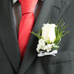 Pinned neatly to the breast pocket of a dark charcoal suit, this classic boutonniere showcases a single ivory-white rose, its petals smooth and luminous as though lit by gentle afternoon light drifting over North Watford rooftops. The rose is fully formed yet not overly open, holding the promise of the day close at its centre. Around it, clusters of baby's breath gather, their tiny white blossoms forming a delicate halo that softens the transition from flower to fabric. Wisps of fern fronds and looping strands of glossy greenery extend outward, adding a natural, slightly untamed note that recalls a quiet walk along the Grand Union Canal. Beneath the bloom, a narrow white ribbon is tied into a crisp bow, the satin edges clean and measured, suggesting a thoughtful, almost ceremonial preparation. The charcoal suit fabric provides a rich, textured backdrop, making every petal and leaf edge stand out with quiet clarity. There is an air of polished restraint here: nothing is excessive, yet each element feels deeply intentional, chosen to mark a milestone with grace. You can imagine the wearer adjusting the lapel one last time before stepping into a wedding venue or formal gathering in North Watford, feeling the cool touch of the ribbon and the subtle weight of the rose. This small floral accent encapsulates tenderness, tradition, and the poised emotion that surrounds life's most meaningful occasions.