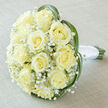 Soft, luminous cream roses gather into an elegant, rounded bridal bouquet, photographed from above so the full, domed composition is clear and immediate. Approximately fifteen to twenty pale ivory roses compose the heart, their petals unfolding in precise spirals that read as both classic and tender; the subtle buttery undertone of each bloom catches the light and imparts a warm, comforting glow. Interspersed among the roses are fine clusters of white Gypsophila, their tiny blooms forming a feathery, cloud-like matrix that lightens the visual weight and imparts an airy, romantic texture. Slim green blades - likely lily or bear grass - sweep and curve around the perimeter, creating a modern heart-like frame that lifts the roses and adds elegant movement. At the base, natural stems peek from beneath a pristine satin wrap: smooth white ribbon is tightly bound around the grip, finished with a neat knot that speaks of careful hand-tying. The bouquet rests on a pale, off-white linen cloth with a very soft weave; the diffuse daylight in the scene enhances the roses' creamy hues, accentuating petal depth and the gentle sheen of the satin. I imagine delivering this to a bride stepping out from a historic terrace in North Watford or pausing near Watford High Street before heading to a small family ceremony - a design that feels both refined and accessible, carrying the scents of soft roses and green grass and offering a quiet, joyful elegance for weddings, anniversaries, or intimate vows.