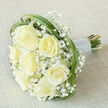 An elegant bridal bouquet photographed diagonally on a neutral beige linen, composed of seven prominent, creamy-white roses arranged with considered symmetry and tender care. Each rose is at a different stage of bloom, petals unfurling in buttery, satin-like folds that reflect the soft, even studio light. Nestled among them are frothy clusters of baby's breath (Gypsophila), their tiny white blossoms creating a weightless, ethereal veil that softens the roses' structure and introduces a whisper of innocence. Arcing loops of vivid green bear grass are woven around the bouquet's edge, their sleek, architectural lines framing the flowers and offering a crisp, contemporary contrast to the romantic whites. The stems are bound into a firm handle and swathed in a silvery-white satin ribbon; along the ribbon, multiple rows of small, luminous pearl beads are fastened with care, supplying a delicate, heirloom-quality accent that glints like dew. The arrangement carries a subtle, fresh fragrance - light rose with a green lift - and the texture ranges from the plush rose petals to the feathery gypsophila and the clean, almost cool touch of the ribbon. As a local florist familiar with North Watford's weddings and quiet ceremonies around Watford High Street and the Vicarage Road area, I imagine this bouquet being chosen for brides who value timeless refinement. This design is available for local delivery from Flower Delivery North Watford and is composed to read beautifully in photographs and in the bride's hands alike.