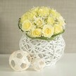 A stylish, contemporary floral display fills the scene, centered on a tight dome of light yellow roses whose petals open generously, creating a plush, almost quilted surface of creamy colour. Between these roses, fine clusters of white baby's breath weave in and out, adding a frothy lightness that softens the overall look and suggests the airy charm of early spring. At the base, a neat band of smooth green foliage encircles the bouquet like a ribbon of fresh leaves, providing a crisp contrast to the pale tones above. The arrangement is nestled into a striking white woven vase, where slender strands cross and loop to form an open, basket-like lattice; through its gaps, hints of shadow and background colour create a sense of depth and lightness. The vase rests on a pristine white surface that reflects a subtle glow, as though set on a carefully laid wedding table in a North Watford venue, ready for guests arriving from nearby roads and parks. Just to the side, two decorative spheres made from pale, natural rattan-style strips echo the same woven motif, one larger, one smaller, arranged casually yet thoughtfully. The backdrop is a soft, blurred beige that could easily be the soothing wall of a North Watford townhouse, keeping every focus on the flowers, textures, and interplay of light. Together, these elements create a centerpiece that feels modern yet romantic, perfect for wedding receptions, engagement dinners, or intimate celebrations where understated elegance and natural materials are appreciated.
