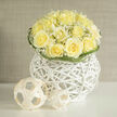 There is a quiet, refined harmony to this arrangement: creamy yellow roses gathered into a near-perfect dome, their ruffled centres unfurling with soft, buttery warmth, encircled by clouds of tiny white gypsophila that breathe lightness between each bloom. The flowers sit within a modern white wicker orb-an airy, architectural vessel whose crisp, sculptural lines lift the composition and introduce a tactile contrast between smooth petals and woven texture. Subtle loops of deep green foliage arch gently around the rim, adding a rhythmic frame and a graphic counterpoint to the bouquet's softness. Scattered nearby are minimal woven spheres that add a contemporary, artistic accent and a hint of whimsy without distracting from the blooms. Under natural daylight the roses take on a luminous quality; their scent reads like sun-washed linen, mingling pure rose notes with an earthy, meadow-like undertone that recalls early summer mornings. As a local florist working in North Watford, I picture this arrangement brightening a mantelpiece near North Watford station or arriving as a thoughtful congratulatory gift for a neighbour from Watford High Street; its restrained elegance is equally fitting for an understated wedding table or a heartfelt message of gratitude. Crafted with careful balance and attention to proportion, the bouquet demonstrates the steady eye and gentle hands of experienced floristry: simple materials, tastefully arranged to produce a composed, hopeful statement that feels both modern and timeless. Available for local delivery across North Watford, it embodies the calm, considered style many of my clients prefer.