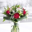 A luxurious red and white bouquet stands proudly in a clear cylindrical glass vase, its fresh green stems visible beneath the waterline like a neat, living framework. At the heart of the design, velvety deep red roses open in gentle spirals, their petals soft and romantic, echoing classic love stories shared in homes around North Watford. A large, intricately ruffled crimson carnation anchors the front of the arrangement, its frilled petals creating rich texture and drama. Around these bold red blooms, clusters of creamy and pure white flowers bring contrast and calm: white alstroemeria with fine speckles in their throats, creamy daisy-style blooms, and several closed white lily buds that promise to unfurl over the coming days. Wisps of delicate baby's breath are tucked between the flowers, their tiny white blossoms adding an airy, cloud-like softness that lightens the whole piece. The bouquet is framed by an abundance of greenery, with broad, glossy dark green leaves carefully curved and wrapped to form a structured collar at the base. Eucalyptus-like foliage and pointed leaves in varied shades of green stretch upwards and outwards, adding movement, depth, and a natural garden feel, much like the green corners of nearby Cassiobury Park. Gentle, even daylight falls across the arrangement, likely from a nearby window, making each colour appear crisp and luminous against a simple white tabletop and softly blurred pale background. The overall mood is polished yet warm, ideal for a heartfelt anniversary surprise, a birthday celebration, or a thoughtful delivery to brighten an office or home somewhere along St Albans Road in North Watford, expressing admiration, joy, and sincere affection in one elegant display.