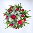 Viewed from directly above against a clean white background, this generous bouquet appears like a vivid, circular tapestry of garden colour, carefully hand-tied by a North Watford florist. Deep crimson red roses form the visual heart, some still in tight bud, others just beginning to unfurl velvety petals that hint at romance and enduring affection. Nestled between them are ruffled carnations in vibrant fuchsia and ruby tones, their plush, frilly blooms creating a sense of movement and celebration. Clusters of small, daisy-like white spray chrysanthemums are scattered throughout, their crisp petals and sunny centres adding brightness and clarity to the mix. Floating delicately around the main flowers, countless tiny white gypsophila blossoms give the impression of soft mist or scattered stars, softening each edge and filling every gap with gentle lightness. The flowers are encircled by a rich collar of foliage in varied shapes and greens: broad, pointed dark leaves, likely from lilies or alstroemeria, provide strong lines and structure, while silvery-green eucalyptus with its rounded leaves weaves between stems, introducing a cool, contemporary tone and a subtle, fresh scent. The natural daylight in the photograph enhances the reds and whites while keeping shadows soft, making the arrangement feel as though it has just been lifted from the workbench of a local shop near Cassiobury Park. The overall impression is one of joy and vitality, perfect for birthdays, anniversaries, or to send warm wishes to someone in North Watford, whether they live near St Albans Road, Watford Junction, or the quieter residential streets, with every petal conveying heartfelt care and thoughtful intention.