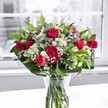 A luxurious bouquet of deep red roses, matching red carnations, white alstroemeria, and clouds of tiny white gypsophila stands in a clear glass vase on a bright white table, bathed in soft natural light from a large window. Each rose, some fully open with velvety petals and others still in bud, draws the eye with its rich crimson tone, while the ruffled carnations add extra fullness and a touch of classic romance. The white alstroemeria, many still closed or just beginning to unfurl, introduce a gentle brightness, their slender petals tipped with the faintest trace of green that echoes the surrounding foliage. Sprays of baby's breath thread delicately between the larger blooms, creating a light, cloudlike haze that softens the stronger reds and makes the whole arrangement feel airy and refined. Around the base, broad, glossy green leaves form a structured collar, holding the flowers in a generous, rounded shape, while finer, more flexible greenery weaves upward, adding movement and depth. The stems are visible through the clear vase, neatly trimmed and submerged in clear water that suggests just-made freshness, like a bouquet that has just arrived from a trusted North Watford florist. Behind, the window view is blurred into gentle greys, so all attention rests on the vivid reds, whites, and greens, much as a thoughtful gift might brighten a quiet living room off St Albans Road. The mood is quietly celebratory and heartfelt, perfect for an anniversary dinner, a romantic gesture delivered across North Watford, or a sincere thank-you arranged to bring colour and warmth into someone's day.