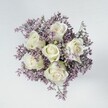 High above a small table in North Watford, the camera looks straight down onto a perfectly rounded bouquet where six creamy white roses sit at the very centre, their petals tightly layered yet beginning to unfurl in gentle spirals. Each bloom is pristine, the velvety petals subtly edged with the faintest hint of pale green at the base, giving them a cool, fresh clarity that suggests purity and quiet devotion. Around them, like a soft halo of mist, lies a dense collar of lilac and muted lavender statice, each tiny flower forming a delicate cloud that frames the roses without overwhelming them. The limonium creates a feathery texture, its myriad miniature blossoms and slender stems weaving together into a cushion of muted purple that deepens the sense of calm and stillness. The arrangement is compact and carefully composed, its circular form bringing a feeling of wholeness, as if the flowers are gathered into a single, comforting embrace. Beneath the bouquet is a smooth, light grey or off-white surface, perhaps a kitchen worktop or coffee table in a North Watford home, its simplicity allowing the gentle contrast between white and lavender to shine. The lighting is soft and even, like daylight filtering through a window on a quiet afternoon, casting only the faintest shadows and highlighting every petal fold and airy sprig. The overall mood is one of understated elegance and serenity, a bouquet suited to moments of reflection, sympathy, or tender celebration, whether placed beside a bed, on a small dining table, or delivered as a thoughtful gesture across the neighbourhoods surrounding St Albans Road.