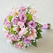 A hand-tied bridal-style bouquet is photographed from above on a soft, light beige fabric, allowing every flower to stand out clearly. At the centre, full lavender and mauve roses open in layered spirals, their velvety petals suggesting calm romance and gentle luxury. Threaded between them are airy stems of pink Alstroemeria, each trumpet-shaped bloom brushed with yellow at the throat and speckled with fine maroon freckles, like tiny brushstrokes. Bright lime-green button chrysanthemums form neat, rounded domes, their tightly packed, almost fuzzy florets adding playful texture and a modern twist to the pastel palette. Cloudlike sprigs of white gypsophila drift through the arrangement, softening edges and giving the bouquet a light, almost misty quality, as though it has just been carried through Cassiobury Park on an early spring morning. Deep green leaves cradle the flowers from beneath, offering a lush frame and subtle contrast. The stems are neatly gathered and wrapped in a smooth fuchsia satin ribbon, finished with a single line of small pearl beads that catch the light in an understated shimmer. Everything about the bouquet feels intimate yet celebratory, perfect for a North Watford bride walking into a local church, an anniversary dinner at home, or a thoughtful surprise delivery across the neighbourhood. The overall impression is of freshness, craftsmanship, and quiet joy, a carefully composed posy that blends classic romance with a touch of contemporary colour and texture, ready to be held in both hands and admired up close.
