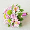 A softly lit, hand-tied bouquet rests against a neutral, linen-like background, its colours glowing gently as if under the light that streams through a North Watford townhouse window. At its heart, several pale pink roses unfold in classic spirals, their petals silky and full, suggesting romance and calm confidence. Around them, playful lime-green button chrysanthemums add a bright, contemporary twist; their rounded, textured heads create visual movement, like little bursts of fresh energy among the calmer tones. Pink alstroemeria weave through the design, each petal etched with fine raspberry streaks and warm, golden centres, giving an impression of delicate artistry. Fine sprays of white baby's breath are tucked in between, softening each transition and creating a light, cloud-like veil over the more defined shapes. Encircling the blooms, glossy green foliage adds depth and a garden-fresh feeling, reminiscent of a morning walk past the trees near North Watford's quiet residential streets. The stems are gathered neatly and bound with a rich fuchsia satin ribbon, which glints slightly in the soft lighting and pulls the pink palette together with a touch of elegance. The overall mood of the bouquet is hopeful and tender, making it easy to imagine it being carried down the aisle, offered as a proposal bouquet, or delivered as a surprise to a doorstep in North Watford. Each carefully chosen flower and the balanced composition reveal the steady hand and practiced eye of a local florist who understands both modern style and enduring romance.