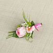 This image shows a meticulously crafted pink rose boutonniere laid on a softly woven fabric, the kind of finishing touch you might choose for a North Watford wedding or prom celebration. The central rose is fully formed yet still tender, its petals unfolding in a soft spiral from a cool, deeper pink core to paler, almost glass-like edges that catch the light. Tucked alongside, delicate alstroemeria petals in blush pink cradle the rose, each bloom marked with faint streaks and a golden-yellow centre that draws the eye inward. Sprays of white gypsophila dot the arrangement like a scattering of tiny pearls, adding airiness and contrast against the richer tones. Slender strands of greenery-some fine and feathery, others broader and gently curled-fan out with poised movement, giving the boutonniere an organic energy while keeping the look polished. The stems are firmly bound with a glossy pink ribbon, wrapped with the precision of a jeweller, its smooth sheen echoing the satin lapels of a suit jacket worn at a reception near Garston or along St Albans Road. The texture play is tangible: velvety rose petals, crisp alstroemeria, cloud-soft gypsophila, and the subtle ribbing of the fabric beneath. The overall mood is one of thoughtful romance and understated luxury, as though an experienced local florist at Flower Delivery North Watford has composed a small floral poem to be worn over the heart. It suggests fresh, early-morning preparation, same-day delivery timed to arrive just before the photographer knocks, ready to mark a day of joy, commitment, and new beginnings.