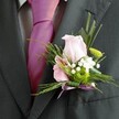 There's an inviting elegance woven through this North Watford buttonhole, as captured in a close-up view of a formal lapel tenderly adorned for a day of celebration. The luminous pink rose takes centre stage-its petals soft, velvety, and open just enough to evoke the anticipation of joyous occasions, much like the first blooms in springtime along Watford's tree-lined avenues. Nearby, a miniature lily reveals vivid, painterly stripes deep within its pastel petals, a flourish echoed in the confident pop of fuchsia from the tie cascading down the shirt's centre. The boutonniere hums with gentle contrasts: crisp green button chrysanthemums sit against clusters of white gypsophila, all cushioned by feathery fronds and slender blades of dark green foliage that ripple with subtle movement. The polished lavender satin ribbon fastening the arrangement completes the visual poetry-its understated shimmer matching both the colour harmony and the occasion's formality. The suit's charcoal bolts and neatly tailored lines offer a sense of dignified celebration, grounding the fresh, romantic colours above. Altogether, this design captures the spirit of weddings and formal events across North Watford: a harmonious celebration of new chapters, crafted with an artist's eye and a local's knowledge of what makes every moment truly memorable.