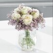 A close-up photograph shows a graceful bouquet of creamy white roses rising from a soft haze of lavender-purple statice, arranged with the calm precision of an experienced North Watford florist. Each rose is fully formed yet still fresh, its velvety petals gently unfurling to reveal a faint blush of pale yellow at the centre, suggesting that the blooms were cut only hours ago, perhaps from a nearby Hertfordshire grower. Around them, countless tiny statice blossoms create a cloud of colour, their papery, clustered petals adding texture and a subtle countryside charm, reminiscent of walks through Cassiobury Park when the wildflowers are in bloom. The stems of both the roses and statice gather neatly into a clear glass vase with a distinctive crackled, ice-like finish; the textured glass catches the natural daylight and scatters it in fine glints, so the entire arrangement seems to shimmer gently on its bright white tabletop. Through the transparent water inside the vase, the strong green stems are clearly visible, lined up in a tidy spiral that reveals the florist's practiced hand. The surface beneath is clean and glossy, reflecting a faint halo of light and casting only the softest shadow, while the background is blurred into a pale grey, keeping every ounce of attention on the flowers themselves. The overall mood is one of quiet reassurance and understated elegance, as if this arrangement has been set down on a bedside table in a North Watford home to comfort someone recovering, or placed on a simple desk to bring a sense of serenity to the working day. It feels ideal for a thoughtful thinking of you gesture, a sympathy gift, or a peaceful anniversary surprise delivered locally with care.