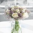A full, round bouquet of creamy white roses and delicate lavender statice stands in the centre of the image, arranged in a tall, clear cylindrical glass vase that rests on a bright white surface. Around a dozen white roses form the heart of the display, each bloom at that perfect stage between bud and full flower, with velvety petals gently unfurling to reveal softly swirling centres. Their colour sits between ivory and warm cream, giving the arrangement a refined, timeless elegance. Threaded between the roses, clouds of tiny, pale lavender-purple statice create a soft, misty halo, their papery, clustered flowers adding a fine, lacy texture that contrasts with the smooth, plush rose petals. The stems are clearly visible through the water-filled vase, upright and freshly cut, their vivid green tones echoing the freshness of a bouquet that might have just arrived from a North Watford florist. Natural daylight appears to pour in from an unseen window, diffusing across the background and leaving it gently blurred, so all attention remains on the blooms. The light picks up subtle shadows and highlights on each petal, giving the roses a luminous glow and catching the occasional glint on the water line in the vase. The scene feels quiet, calm, and hopeful, like a peaceful corner of a North Watford townhouse near Cassiobury Park or a sunlit reception desk along St Albans Road. This arrangement would suit many tender moments, from a thoughtful sympathy gesture to a graceful anniversary surprise, bringing a sense of understated comfort and classic beauty to any contemporary space.