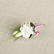 Arranged with the care of an experienced North Watford florist, this boutonniere rests on a softly textured, light woven fabric, as though just placed down before being pinned to a lapel. At its heart, a single white lisianthus bloom opens fully, petals curling and rippling like soft cream, giving a sense of gentle luxury without ever feeling showy. Tucked snuggly at its side are two lisianthus buds, slightly creamy at the edges, still wrapped in themselves and suggesting new beginnings, much like a wedding morning in the quiet streets near Cassiobury or along St Albans Road. Wispy sprays of baby's breath peek through the design, their tiny, cloud-like flowers scattered delicately between the blooms, adding a snow-dust lightness and a dreamy, romantic haze. Slender, reed-like greenery weaves around and behind, its elegant lines adding a touch of movement, so the boutonniere never feels stiff, but rather alive and freshly gathered. Anchoring the composition is a band of vivid pink satin ribbon wrapped carefully around the stems; its polished, satiny sheen glows gently and is tied into a neat bow that brings just the right amount of playful charm. The colour contrast between the bright ribbon and the tranquil whites and greens creates a modern yet timeless look. Set against the neutral woven backdrop, every texture is heightened-the silkiness of petals, the airy baby's breath, the smooth ribbon finish-making this piece an ideal choice for grooms, ushers, or prom guests around North Watford who want something refined, personal, and quietly memorable.