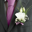 A wedding boutonnière, carefully pinned to a charcoal suit lapel, brings a soft, romantic glow to the scene, as if capturing the quiet excitement of a North Watford ceremony. At its centre is a flawless white lisianthus bloom, the petals layered in ruffled rings that look almost translucent at the edges, like fine silk lit from within. Behind the main flower, two slim lisianthus buds rise elegantly, still closed, their tapered shapes suggesting new beginnings and the future taking gentle root. Threaded through the composition are feathery sprays of baby's breath, tiny white blossoms scattered on delicate stems, giving the whole piece an airy, almost mist-like halo. Subtle variegated foliage peeks through in slender lines of green, adding organic movement that keeps the boutonnière from feeling too formal, while a single rolled deep green leaf curves beneath the flowers, offering structure and depth. The stems are bound with a mauve ribbon, tied into a neat little bow that brings a muted hint of lavender tone, echoing the groom's tie and adding a contemporary twist to a classic design. The textures are soft and inviting: velvety petals, crisp greenery, and the faint shimmer of satin ribbon, all combining into something intimate rather than showy. You can almost imagine the clean, powdery scent of fresh flowers mingling with the cool air just outside the registry office or church. Crafted with the eye of an experienced local florist, this boutonnière feels like a thoughtful nod to tradition, yet rooted in the relaxed, genuine spirit of weddings celebrated across North Watford.