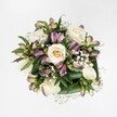 A meticulously arranged bouquet is photographed from directly above, every bloom clearly outlined against a spotless, bright white background that feels like soft daylight filtering into a North Watford kitchen. At the heart of the design, creamy white and soft ivory roses nestle closely together, some still slightly cupped with a hint of pale buttery yellow at their centres, others fully open so their velvety petals spiral outward in generous layers. Threaded between these classic roses are graceful stems of alstroemeria, or Peruvian lilies, their slender buds and partially opened flowers revealing petals washed in mauve, lavender, and purplish-pink, gently brushed with green along the outer edges and throats. Tiny star-like blossoms of pure white gypsophila float between the larger flowers, creating a delicate, cloud-like veil that softens every transition and brings a sense of weightless romance. A collar of rich, deep green foliage forms a sturdy base, the broad leaves glossy, folded, and slightly curved as if carefully wrapped by an experienced local florist preparing an order for delivery near Garston or along St Albans Road. The greens anchor the bouquet and emphasize its freshness, framing the blooms in a way that looks both natural and expertly composed. The overall impression is of calm, polished beauty-an arrangement that could just as easily be placed on a wedding table at a North Watford venue as on a bedside to offer quiet comfort. One can almost imagine the faint, clean fragrance of the roses and the subtle rustle of leaves as the bouquet is gently turned in the florist's hands, showcasing the artistry behind every stem and the thoughtfulness of sending such flowers to mark love, gratitude, or remembrance.