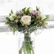 An elegant white and green bouquet stands in a clear cylindrical glass vase, placed before a softly glowing white background that feels like daylight streaming through a North Watford window. Three creamy white roses form the calm, central focus, their petals gently unfurling in layered spirals, each bloom flawless and velvety, with subtle ivory tones at the heart suggesting purity and new beginnings. Around them, muted mauve and blush alstroemeria buds and blossoms weave through the design, some still half-closed, others just opening, their slender petals streaked with delicate markings that add quiet interest rather than bold colour. Wisps of white gypsophila, or baby's breath, thread through the arrangement like tiny clouds, their miniature blooms resting on fine green stems that drift above and between the larger flowers, softening every edge. Broad, folded emerald leaves and smaller sprays of lush foliage cradle the roses and alstroemeria, giving the bouquet a rounded, compact shape with a polished, professional finish. Through the vase's transparent glass, the green stems can be seen gathered neatly and tied with a subtle ribbon just above the clear waterline, evidence of a careful florist's hand. The overall mood is peaceful and refined, as if the bouquet were prepared early in the morning for a quiet moment of reflection in a Watford home. It feels suited to sending comfort, expressing sympathy, or marking a thoughtful milestone, the gentle play of white and green echoing the calm of nearby Cassiobury Park on a still day, while the soft lighting wraps everything in a serene, almost ethereal glow.